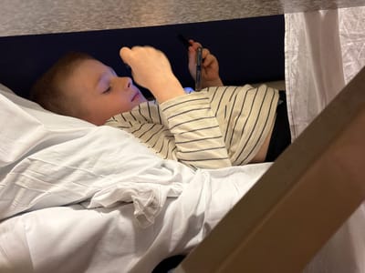 A young child lies in a bed with white linens, possibly in a bunk bed or confined space, as indicated by the close proximity of the overhead structure. The child is looking at and holding a mobile phone with a protective case. The child is wearing a striped, long-sleeved shirt. The location is not discernible as being Kamensk-Shakhtinsky, Russia, nor any city without further context.
