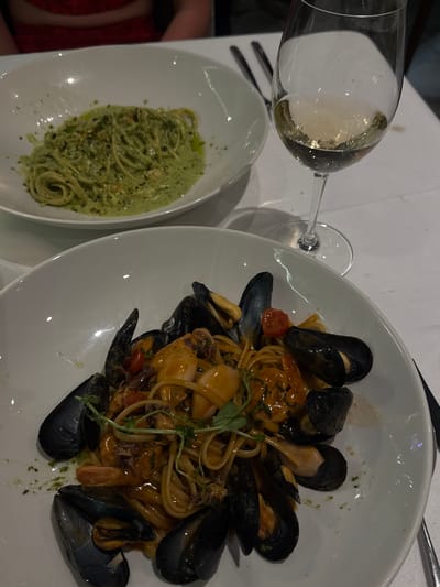 In a restaurant setting in Saint Paul's Bay, Malta, two plates of pasta are presented on a white tablecloth. In the foreground, a white bowl contains spaghetti with mussels, calamari, and shrimp in a light orange sauce, garnished with microgreens. A glass of white wine is positioned to the right of this plate. Behind it, another white bowl holds spaghetti with a green pesto sauce, sprinkled with chopped nuts. The image is a close-up shot of the meals, with a blurred background showing the indistinct torso of a person wearing a red garment.