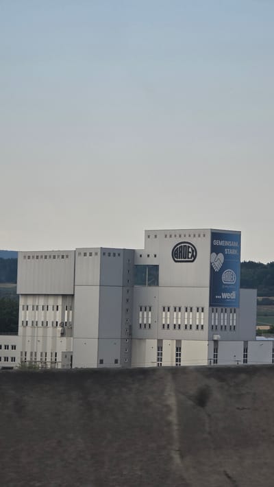 The image shows a tall, modern warehouse or industrial building in Loosdorf, Austria. The building is primarily gray and white, with numerous windows arranged in rows. A large blue banner is prominently displayed on the upper part of the building, featuring text in German that reads "GEMEINSAM. STARK." (Together. Strong.) and includes logos for "ARDEX" and "... wedi". The Ardex logo is also visible in a large oval on another part of the building. The sky above is overcast and light gray. To the right of the building, a dark, textured surface, possibly asphalt or gravel, dominates the foreground, suggesting an industrial yard or parking area. The scene appears to be set during the daytime under a cloudy sky. There are no people visible in the image, and the overall impression is one of industrial activity and business. The architectural style suggests a functional, large-scale facility.