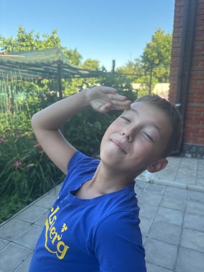 A young boy, with his eyes closed and a faint smile, salutes the camera with his right hand raised to his forehead. He is wearing a blue t-shirt with yellow lettering that appears to be a logo. The background suggests an outdoor setting, possibly a backyard, with greenery and some sort of netting structure visible. The boy's gesture and closed eyes might indicate a playful or humorous moment. The overall lighting suggests it is daytime, likely sunny. The scene appears to be in Zmiiv, Ukraine.