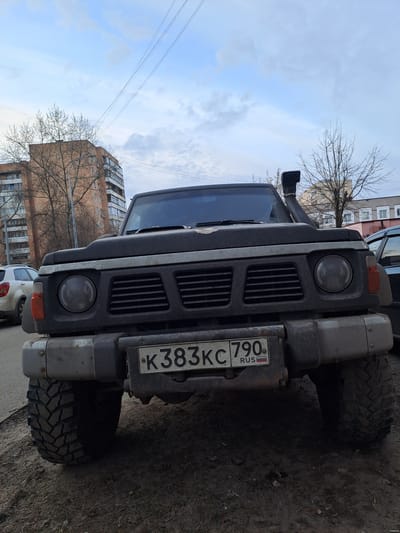 A dark-colored, possibly black or dark gray, off-road vehicle, likely a Nissan Patrol, is depicted in a front-facing, low-angle shot on a dirt and mud surface in Yegoryevsk, Moscow Oblast, Russia. The vehicle is equipped with a heavy-duty grey front bumper, round dark-tinted headlights, and a snorkel attached to the passenger side. Its front license plate displays "K383KC 790 RUS." The paintwork on the hood appears worn. Large, aggressive-tread off-road tires are visible. In the background, a multi-story brick apartment building is present to the left, partially obscured by bare trees. A lighter-colored vehicle, possibly a white SUV, is parked behind the main subject on the left. To the right, another building and additional bare trees are visible. The sky is overcast with patches of lighter blue, and utility lines stretch across the upper portion of the frame.