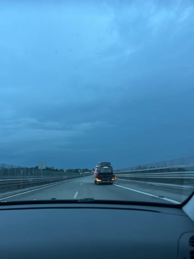 The image depicts a view from inside a vehicle traveling on a highway in Novi Han, Bulgaria. The perspective is from the passenger seat, looking out the side window and windshield. The highway stretches ahead, with multiple lanes delineated by white lines. The sky is overcast and a deep blue, suggesting it is either early morning or late evening, or that the weather is gloomy.

Several vehicles are visible on the road. In the foreground, a black truck is prominently featured, its brake lights illuminated, indicating it might be slowing down or stopped. Further down the highway, other vehicles can be seen as specks of light, suggesting a moderate amount of traffic. The sides of the highway are lined with a concrete barrier and vegetation, including trees.

A noticeable detail is the reflection of the interior of the vehicle on the window, particularly the dashboard and part of the driver's side. The image is oriented vertically, with the highway appearing to run from the bottom left to the top right of the frame, and the sky dominating the left side. There is no visible text in the image. The overall mood is serene, with the vast sky and the steady movement of traffic on the highway.