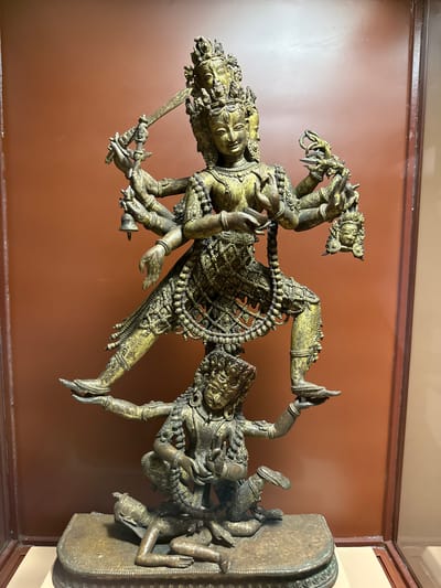 The image shows a bronze sculpture of the Hindu deity Kali trampling Shiva. Kali is depicted with multiple arms, holding a sword and other objects. She is standing on Shiva, who is lying on a pedestal. The sculpture is on display at the Patan Museum in Lalitpur, Nepal. The backdrop is a plain brown wall.