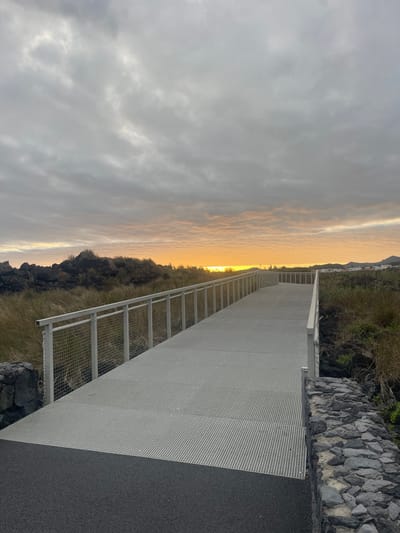 The image depicts a modern, metal walkway stretching out towards a horizon illuminated by a bright, orange-yellow glow, likely at sunset or sunrise. The scene is dominated by the walkway's gray, perforated surface and its safety railings. To the right of the walkway is a dark asphalt surface and a stone wall. On the left is rough terrain covered with dry grass and dark volcanic rock, transitioning into the bright horizon. The sky above is covered in textured gray clouds. Based on the description, this likely depicts the "Ponta do Rosário" viewpoint in Lagoa (Nossa Senhora do Rosário), Portugal, a known location with a similar walkway and scenic outlook. There are no people visible in the image.