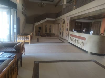 The image shows the reception area of a hotel, likely located in Khiva, Uzbekistan. The reception desk is prominent, with the word "RECEPTION" visible. There are decorative plates on the wall behind the desk. To the left of the reception desk, a staircase leads to an upper level. Further to the left, there is a seating area with wooden furniture, including a sofa and a coffee table with magazines. In the background, there are more seating areas with wooden daybeds. The floor is tiled with a patterned inlay. The overall environment appears to be a well-maintained indoor space, possibly part of a hotel or a similar establishment. A person is partially visible behind the reception desk on the right side of the frame.