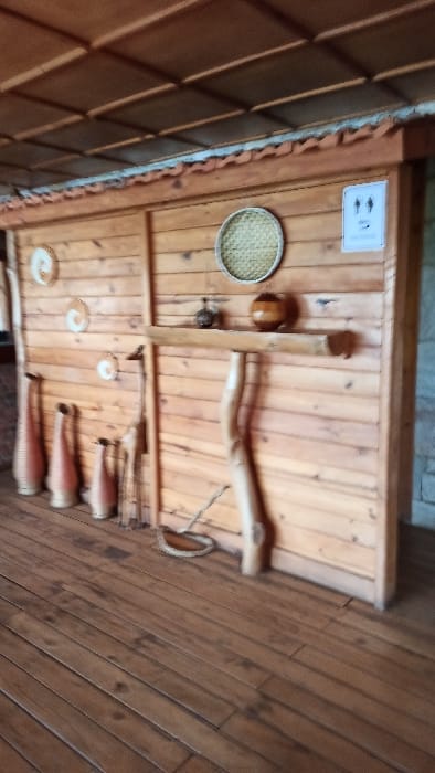 The image displays a rustic interior or sheltered space in Bwishyura, Rwanda, adorned with traditional wooden and ceramic elements. The primary surface is made of warm-toned, light brown wooden planks, likely a wall or ceiling, running horizontally across the frame (when correctly oriented).
A prominent feature is a round, flat woven tray or sieve, crafted from natural fibers, positioned in the upper left section. Nearby, a small, dark, spherical wooden object is attached to the planks. Several pieces of natural, irregularly shaped wooden branches are artistically arranged. A large, curved branch extends across the top-middle, while other smaller wooden pieces are integrated into the display, suggesting a decorative purpose.
On the right side of the wooden surface, a row of at least three elongated, reddish-brown unglazed ceramic vessels, resembling traditional Rwandan pottery such as *ibibindi*, are hung vertically. Further down, several smaller, round, light-colored objects, possibly decorative plates or bowls, are also affixed to the wood.
In the upper left, a small, white rectangular sign is visible, displaying two indistinct, stylized dark figures or icons, too blurry to read any specific text or meaning.
To the far left of the main wooden display, a section of terracotta-colored, possibly corrugated roof tiles or an architectural overhang is visible, suggesting an open-sided structure or a view towards an exterior element.
No human activity is depicted; the scene is a static cultural display. The lighting appears bright and natural, indicating a daytime setting. The arrangement of crafts evokes a sense of traditional Rwandan artistry and craftsmanship.