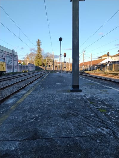 An eye-level shot shows a train station platform extending into the distance. Train tracks run parallel on either side of the platform, converging towards the horizon. Overhead wires crisscross the sky above the tracks. A large, gray, square support post is near the foreground. A signal light post stands on the platform further away. A building with a patterned pink facade is partially visible on the left, and a long, one-story building lines the tracks on the right. The platform surface is dark with patches of lighter gray. The sky is blue, and the lighting is bright.