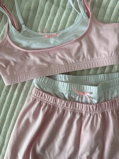 A close-up, overhead shot shows a pink and white pajama set with a textured, off-white blanket. The pajama set includes a camisole-style top and matching shorts. Both pieces are made of a soft, cotton-like material. The top is pink with white straps, and features a small pink bow at the center of the neckline. The shorts are primarily pink with a white waistband, also adorned with a pink bow. The blanket beneath the pajamas has a ridged texture, creating a visual pattern of raised lines. The scene is well-lit, with soft shadows indicating diffused lighting, possibly from a window or overhead light source. The image does not depict any specific location in Ivatsevichy, Belarus, nor does it contain any people or actions.