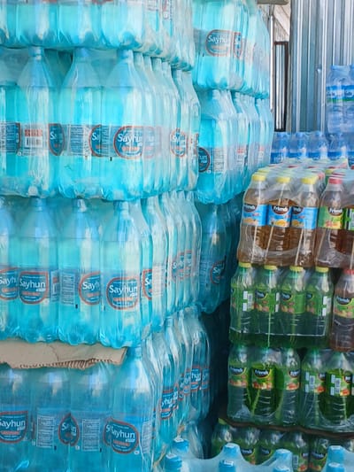 The image shows a large collection of bottled water and beverages stacked in an outdoor area, likely a retail or storage space. The bottles are predominantly clear plastic and contain a light blue liquid, consistent with bottled water. Many of the bottles are wrapped in clear plastic packaging, and some also have printed labels with the brand name "Sayhun".

In the right portion of the image, there are stacks of bottles containing beverages with different colored liquids, including a light brown and a green liquid. These bottles also feature prominent labels, with one visible brand name being "Friends". The stacking appears organized, with some bottles in larger multi-pack shrink wrap.

The background of the image includes elements that suggest an urban or commercial setting. To the right, there is a structure with corrugated metal siding, possibly a building wall or a temporary shelter. The ground surface is not clearly visible. The lighting suggests it is daytime.

The image is taken from a slightly elevated angle, looking down at the stacked items. The overall impression is one of a commercial inventory of bottled drinks. There are no people visible in the image, and no specific actions are depicted beyond the arrangement of the products. Based on the presence of the "Sayhun" brand, which is a well-known water brand in Uzbekistan, and the architectural cues, this scene is likely located in the city of Gulistan, Uzbekistan.