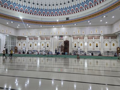 The image shows the interior of a mosque, likely located in Paya Tringgadeng, Indonesia, given the context provided. The mosque features a large, domed ceiling adorned with intricate patterns in blue, red, and gold. A decorative frieze with Arabic calligraphy runs along the upper walls. The walls themselves are elaborately decorated with geometric patterns and arched niches.

In the foreground, the polished tiled floor reflects the overhead lights and the details of the mosque. Several individuals are present, most of whom are seated on green prayer mats, facing the front of the mosque. The individuals appear to be engaged in prayer or quiet reflection. Two individuals in the left mid-ground are standing and appear to be interacting. In the far mid-ground, near the center, is a wooden minbar (pulpit). A digital clock displaying red and green numbers is visible on the wall. The environment is clean and well-lit, suggesting a place of worship that is maintained with care.