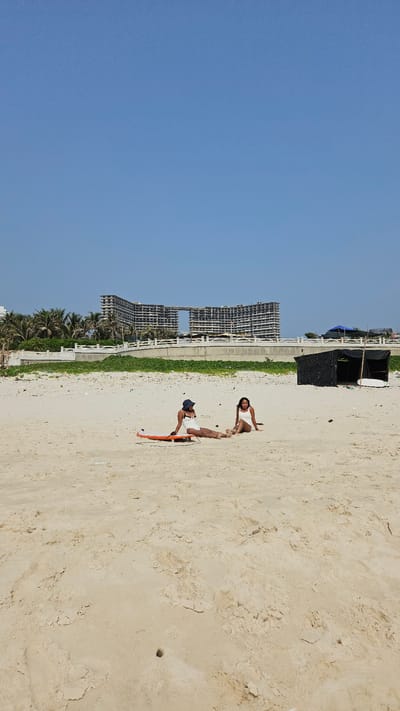 The image captures a sunny day on a wide, sandy beach in Bac Cam Ranh, Vietnam, under a clear, bright blue sky. Two individuals are visible on the light-colored sand. In the foreground, closer to the viewer, a person wearing a dark cap, a light-colored top, and shorts is lying prone on a vibrant orange and white bodyboard or small surfboard. Further back, another person dressed in a light-colored top and shorts is seated directly on the sand.

Behind the beachgoers, there is a small black tent-like structure with a white board, possibly a surfboard or paddleboard, leaning against it. A narrow strip of green vegetation runs along the edge of the beach, separating it from a concrete walkway or barrier. Beyond this, several large, multi-story buildings, characterized by their visible concrete frameworks and lack of finished facades, line the distant horizon, suggesting they are either under construction or abandoned. Palm trees are visible beneath these structures, contributing to the tropical coastal setting. The bright, clear lighting indicates it is daytime, likely midday or early afternoon. The activity appears to be leisurely relaxation on the beach, possibly involving water sports. No visible text is present in the image.