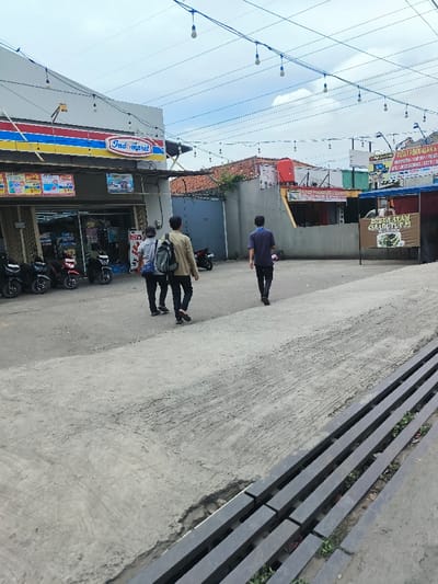The image captures a daytime scene of a commercial area, likely in Indonesia, featuring a convenience store and several people. The sky is overcast with white clouds, suggesting a cloudy day but with ample natural light.
In the foreground, the ground is paved with concrete, displaying visible tire marks or textured patterns. Towards the bottom right, a section of a dark-colored metal grate or drainage cover is visible, with sparse green vegetation growing underneath it.
The midground is dominated by an "Indomaret" convenience store on the left, easily identifiable by its prominent blue, red, and yellow logo with white lettering. In front of the store, several motorcycles or scooters are parked. Through the open entrance of the Indomaret, various products on shelves and in coolers can be seen. Above the entrance, multiple promotional posters with images and pricing information are displayed.
Three young men are walking away from the viewer, heading towards the background. The person on the left wears a light-colored top and dark pants, carrying a light backpack. The middle person has a light brown or beige jacket or shirt, dark pants, and a dark backpack. The person on the right is dressed in a dark blue or black t-shirt and dark pants. They appear to be casually walking through the area.
To the right of the walking men, a brown banner is partially visible, featuring white text that reads "BOBUR AVAY" and "CILADUTUT 29", suggesting a food stall, possibly selling chicken porridge. Further into the background, a red water tank sits atop a building. Another sign on a building in the background displays fragments of text: "PUSAT PEMJUALAN &" and "A/S/GET/S".
Overhead, blue cables with numerous small, unlit light bulbs are strung across the scene, likely for decorative lighting or illumination during the evening. The overall setting appears to be a typical street or commercial complex in an urban or suburban Indonesian environment.