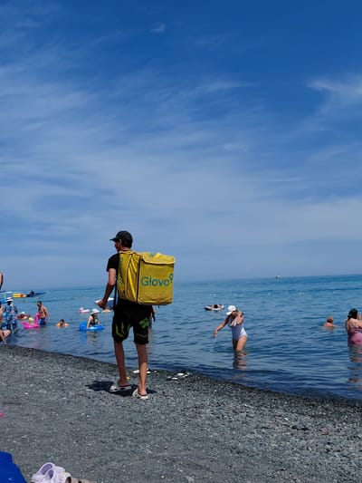The image depicts a beach scene in the city of Ақши, Kazakhstan, on a sunny day with a clear blue sky. In the foreground, a person wearing a black cap and black shorts with green accents is standing on a pebble beach, facing away from the camera. They are carrying a large, bright yellow insulated backpack on their back with the word "Glovo" clearly visible in blue lettering. The person is wearing white slides. In the shallow water, numerous people are enjoying the beach, with some swimming and others wading. Inflatable toys, including a pink flamingo, are visible in the water. Further out in the ocean, several boats are present. The overall scene conveys a typical summer day at the beach with recreational activities.