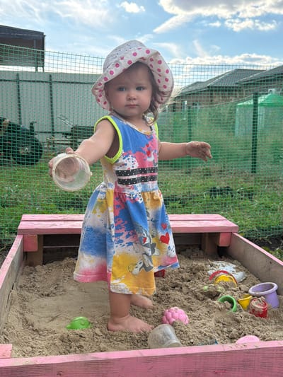 A young child, a girl, is playing in a sandbox. She is wearing a colorful dress with cartoon characters and a polka-dot hat. The sandbox is made of pink wood and is filled with sand and various toys, including buckets and molds. The child is holding a small plastic cup in her hand, and her feet are visible in the sand. The scene appears to be outdoors, with a green fence and a cloudy sky in the background. The overall impression is one of playful innocence and childhood enjoyment. The time of day is likely daytime, given the bright sky. The location is noted as Krasnyi Krym, Russia.