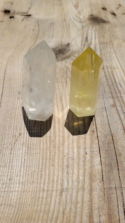 The image shows two faceted crystals, one clear and one yellow, lying on a weathered wooden surface. The clear crystal is on the left, and the yellow crystal is on the right. Both crystals are oriented vertically, with their pointed ends facing upwards. The wooden surface has prominent grain lines and appears to be outdoors, with sunlight casting shadows from the crystals onto the wood. The location of Terrebonne, Canada, is not depicted in the image.