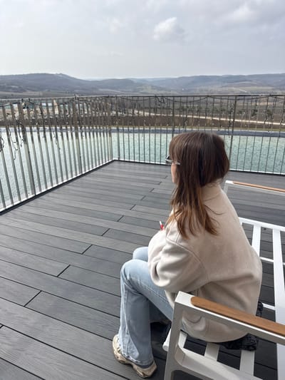 An elevated outdoor deck, likely located in Зеленогорское сельское поселение, Ukraine, is depicted under an overcast sky. A lone female subject, with medium-length brown hair, is seated in a white metal chair with wooden armrests, facing away from the camera and towards the distant horizon. She wears light-colored sunglasses, a light beige/tan collared jacket, light blue jeans, and light-colored athletic shoes. Her right hand holds a red cylindrical object, possibly a pen. The deck features dark grey composite decking boards. A metallic railing with vertical balusters and draped string lights separates the deck from a large, light blue-green body of water, possibly an infinity pool or pond, which extends towards a distant landscape of rolling hills and sparse vegetation.
