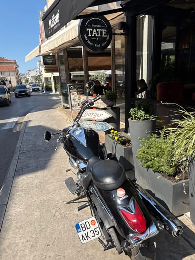A black Yamaha motorcycle is parked on a paved sidewalk next to a building with a large window. The motorcycle has a red taillight and a chrome exhaust pipe. A license plate that reads "BD AK 35 MNE" is attached to the rear fender. Several signs are visible on the building, including one for "TATE Snack Bar Beer Food Stove" and another for "GARDEN CAFE". There are also signs indicating "Burger" and "Pizza". The scene appears to be an outdoor cafe or restaurant. Green plants in planters are near the motorcycle. The lighting suggests it is daytime, likely sunny, with clear blue skies visible. The image is oriented vertically.