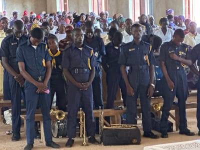 The image depicts a gathering of people, likely in a community hall or church, in Kufana, Nigeria. In the foreground, several young men in matching dark blue uniforms with yellow trim stand attentively. These uniforms appear to be for a marching band or similar organization, as musical instruments, including trumpets and what looks like a drum, are visible at their feet and on a bench. The young men are diverse in complexion and build.

Behind them, a larger group of men, women, and children are visible, many wearing colorful headwraps and traditional attire. Their expressions suggest a solemn or engaged atmosphere. The environment appears to be indoors, with plain walls and what could be wooden benches. The lighting suggests daylight, possibly filtered through windows. The overall scene suggests a community event, possibly a service or ceremony, involving a musical group.