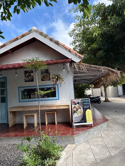 Here's a description of the image:
The image depicts the exterior of a building in Pejukutan, Indonesia. The building has a white facade with blue window and door frames, and a tiled roof. A menu board with the words "Temu Rasa" is visible next to the building, advertising various food items such as Soto, Bakso, and Seafood. A wooden shelf-like structure with cube cutouts is attached to the wall. Greenery is present around the building and in hanging planters. The sky is visible in the background.