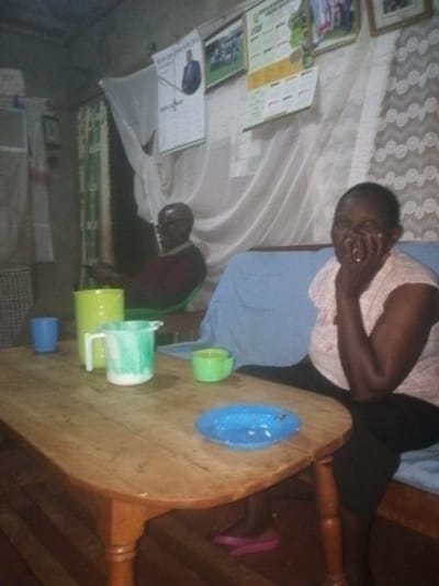 The image captures an intimate, dimly lit indoor scene, likely a living room or common area, featuring two older individuals. A wooden oval-shaped table occupies the foreground and center, laden with various drinking vessels.
On the right side of the frame, an older woman with dark skin and short hair is seated on a blue-cushioned sofa or bench. She wears a light-colored, possibly checkered, short-sleeved top and dark pants. Her right hand is raised to her mouth or jaw, and she has a clear expression of joy, appearing to be smiling or laughing. A ring is visible on her right hand. Her feet, clad in pink flip-flops, are visible under the table.
Further back on the left, behind the table, an older man with dark skin, wearing glasses and a dark, long-sleeved top, sits on a green chair. He is looking downwards or slightly to his left, with his expression not clearly visible. He appears to be engaged with something in his hands.
On the wooden table, from left to right, are a blue plastic cup, a tall bright yellow plastic jug, a light green plastic pitcher (which appears to contain a white liquid like milk), a smaller bright green cup, and a bright blue plastic plate or shallow bowl in the right foreground.
The background shows a wall adorned with several items. To the left of the man, a white curtain or mosquito net hangs. Above the man, a poster features a man in a suit, with the name "DAVID INFANCHO" clearly visible, along with some calendar-like markings below it. To the right of this, other framed images or posters are visible, including one depicting a group of people and another with green and white graphics, possibly a chart or organizational information. A large, light-colored curtain or net drapes across the middle of the background, behind the table and the woman, creating a soft backdrop. Further to the right, a patterned fabric or curtain hangs against the wall.
The lighting is dim, suggesting it is either evening or that the room has limited light sources, contributing to a cozy or modest ambiance. The overall impression is one of a relaxed, domestic setting, possibly during a casual gathering or simply unwinding.