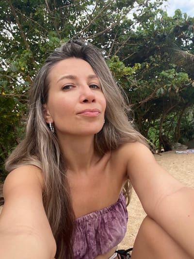 A woman is lying down on a sandy surface outdoors, with green trees and foliage in the background. She is looking up towards the camera with her head tilted. Her face is illuminated by natural light, suggesting it is daytime. She has long, light brown hair with streaks of grey. She is wearing a purple, strapless top with a ruffled neckline. The setting appears to be tropical, possibly a beach or garden area, given the lush greenery and sandy ground. The overall mood is relaxed and casual. There is no visible text in the image.