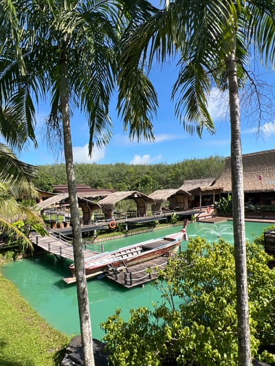 The image depicts a serene scene at a waterfront resort or village, likely in Tambon Pa Kae Bo Hin, Thailand, given the lush tropical vegetation and architecture. The setting is a clear, sunny day, with a bright blue sky and scattered white clouds visible.
The primary subjects are several traditional wooden boats docked alongside a wooden boardwalk and thatched-roof buildings. These buildings appear to be bungalows or shops, constructed with natural materials, suggesting a rustic and eco-friendly aesthetic. The water is a vibrant, emerald green, hinting at its clarity or the presence of algae, and a fountain is visible in the distance, adding to the tranquil atmosphere.
Palm fronds frame the top and left sides of the image, emphasizing the tropical location. The boardwalk is made of weathered wood and leads to the various structures. Life preservers are visible on the boardwalk and near the water, indicating safety precautions.
While no people are clearly visible in the image, the presence of the boats, boardwalk, and buildings implies human activity and habitation. The overall mood is peaceful and relaxing, typical of a tropical getaway. The time of day appears to be midday, judging by the bright sunlight and the shadows cast. There is no visible text in the image.