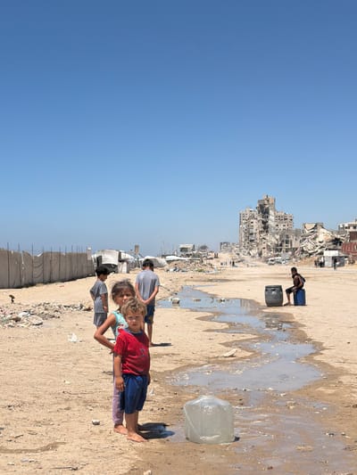 In the city of غزة, Unknown, four children stand on sandy ground littered with debris. The child in the foreground, a boy wearing a red shirt with a Spiderman design and blue shorts, looks directly at the camera. To his left and slightly behind him, a girl with braided hair and a blue and white patterned top stands with her hand on her hip. Behind them, a boy in a gray t-shirt and shorts stands facing away from the camera. Further back and to the left, another boy in a gray t-shirt and shorts is visible. In the background, the remains of bombed-out buildings dominate the skyline under a clear blue sky. A narrow stream of water, likely from a leak or runoff, flows through the sandy terrain. To the right, a young person sits on a blue barrel next to a dark gray barrel. Tents and makeshift shelters are set up on the left side of the image, and piles of rubble are scattered throughout the scene.