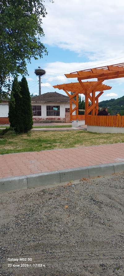 This is a vertically oriented, eye-level shot of a rural scene in Zhaltusha, Bulgaria, likely taken on a sunny day.

The primary subjects are a wooden pergola, a small building with a tiled roof, a stork's nest on a utility pole, a conifer tree, and a large deciduous tree. The pergola, made of light-colored wood, features a slatted roof and is positioned in the foreground, partially obscuring the building. The building itself is white with red-brown trim around the windows, which have metal bars. The stork's nest is a prominent object atop a utility pole to the left of the building, with a stork visible within it. A dark green conifer tree stands in front of the building, and a leafy green tree is on the far left. The ground is covered by a red brick pathway and gravel, with patches of grass.

There is no clear activity or event taking place. The scene appears to be a static view of the environment.

Notable details include the presence of the stork and its nest, suggesting this is a location where storks are common. The presence of a utility pole reinforces the rural setting. The time of day appears to be afternoon, given the quality of light.

Visible text in the lower right corner reads: "Galaxy M52 5G 23 юни 2026 г. 17:03 ч.", which indicates the device used to capture the image, the date (June 23, 2026), and the time (17:03).
