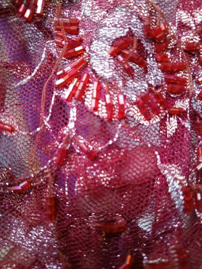 Close-up image showcases a textured textile, primarily colored in shades of red and silver. The fabric features a netting pattern, layered with an ornate design. Red beads are sparsely attached, creating a subtle shimmer. The silver embellishments appear to form floral or leaf-like motifs. The overall aesthetic conveys a sense of luxury and craftsmanship.