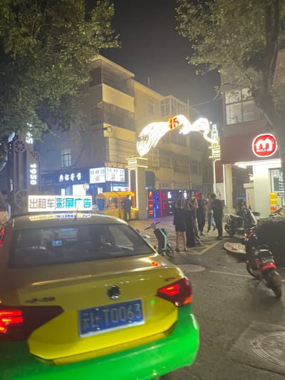 The image captures a nighttime street scene in Dali City, Yunnan Province, China. In the foreground, the rear of a yellow and green taxi is prominently featured, displaying a blue Chinese license plate, "云L•T0063". An illuminated sign on the taxi's roof reads "出租车彩广告" (Taxi Color Advertisement).

Midground elements include a group of approximately eight to ten individuals standing on the patterned sidewalk to the right, some appearing to be engaged in conversation or looking at mobile devices. Parked alongside the sidewalk are several electric scooters or mopeds, including one with a light green frame and another with a red body. Further left, a yellow street food cart is visible with one person standing nearby.

The background consists of multi-story buildings, illuminated by artificial light. A prominent, ornate neon archway spans the street, featuring bright white and orange lights shaped like a large bird or phoenix with outstretched wings, and the number "15". Various commercial establishments line the street level, including a storefront with a large "M.Y.J." sign in red, likely a convenience store, and another with a sign reading "典铂寄售" (Dian Bo Consignment). A tall, narrow sign displaying "1958" is also visible on the left. Overhead, dark tree foliage is visible against the night sky, illuminated by streetlights.