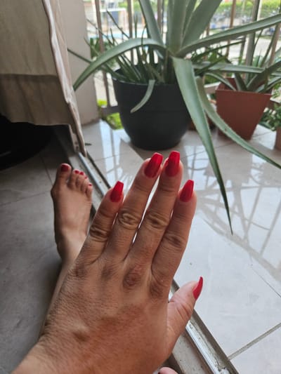 A person's left foot and right hand, both with bright red nail polish, are positioned on a tiled floor near a sliding glass door. In the background, several potted plants are visible, including a large aloe vera plant in a dark pot. The scene appears to be on a balcony or patio in an urban setting. The flooring consists of light-colored rectangular tiles. A portion of a beige curtain is visible to the left of the frame. The lighting suggests it is daytime, with shadows cast by the plants and the structure of the building. The overall impression is one of a relaxed, personal moment captured outdoors.