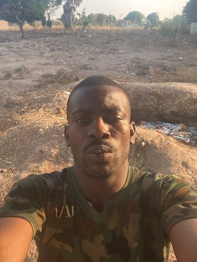 A man in a camouflage shirt takes a selfie outdoors. The background features dry, straw-covered ground, trees, and a mound of trash. Harsh sunlight casts strong shadows. The man's expression is neutral.