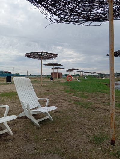 The image depicts a view of a beach or park area on a cloudy day. In the foreground, there are two white plastic beach chairs. One chair is fully visible and positioned to the left, while a portion of another chair is visible to its left. Behind the chairs, there is dry grass and sand. Further in the background, several woven beach umbrellas stand on poles. One of these umbrellas, in the center of the frame, has an orange life preserver hanging from its pole. Beyond the umbrellas, there is a grassy area with more beach chairs and some low wooden structures. In the far distance, there are buildings and power lines, suggesting a developed area or the edge of a town. The sky is overcast with gray clouds. The scene is likely located in Sol-Iletsk, Russia, known for its salt lakes and recreational areas.