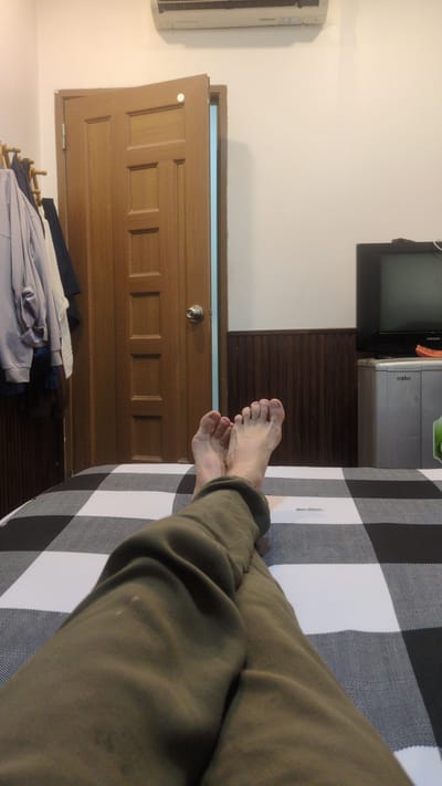 A photograph taken indoors in Phan Rang – Tháp Chàm, Vietnam depicts a pair of bare feet resting on a black and white checkered surface, possibly a bed or blanket. The legs are clad in olive-colored trousers. In the background, a wooden door is slightly ajar, revealing a glimpse of blue light. Adjacent to the door is an air conditioning unit mounted on the wall. Above the door, a retro television set sits atop a metal cabinet. A pile of clothing is visible near the base of the door. The wall behind the television and cabinet is painted white, with a section of wooden paneling below.