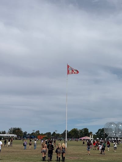 The image depicts an outdoor event taking place on a large, dry grass field in Martínez, Argentina, under an overcast sky. A tall, white flagpole is positioned centrally in the midground, flying a red, white, and blue flag with white text and a logo, consistent with event branding (e.g., "Lollapalooza"). Numerous individuals are distributed across the field, with many walking away from the camera. In the foreground, a group of five people, consisting of four women with long blonde hair and one man, are prominently visible from behind, dressed in dark shorts, crop tops, and boots. In the background, various temporary structures, white tents, stage elements, and stadium-style floodlight towers are visible, interspersed with trees. Two large, translucent, spherical or bubble-like structures are situated on the right side of the frame.
