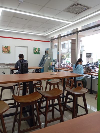 Inside a bright, modern convenience store or cafe seating area, two women are seated at separate wooden tables. The interior features a white ceiling with fluorescent strip lights and a ventilation unit, as well as white walls. A prominent red stripe runs horizontally across the upper section of the left wall.

To the left, a woman with dark hair tied back wears a dark blue or gray hooded top and a black backpack. She is seated at a rectangular wooden table, facing away from the viewer. Next to her on the table is a black helmet. Behind her, against the white wall, are two waste disposal bins labeled "一般垃圾 Trash" and "資源回收 Recycling" in both Traditional Chinese and English. Further back, a white door has text partially visible, possibly reading "請勿靠近" (Please do not approach). An ATM machine is also visible in the background, to the right of the door, with some unreadable text at its top. Two small, framed artworks depicting green, organic motifs decorate the wall above the bins and near the ATM.

To the right, another woman, also with dark hair tied back, wears a light blue hooded top and dark green pants. She sits at a long, wooden bar-style table that runs alongside a large glass window. She appears to be looking down at something on the table or in her lap. Through the window, a street scene unfolds, showing parked motorcycles and scooters, a white car, and multi-story buildings under bright daylight, indicating good weather.

Numerous wooden high stools with curved seats are arranged throughout the seating area, featuring black metal bases. The setting suggests a casual environment for individuals to relax, study, or eat.