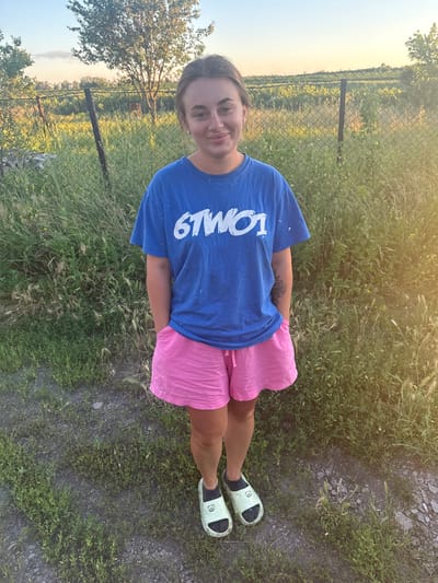 This is a portrait-style, vertical, outdoor, eye-level media capture of a young woman posing in a grassy area with a chain-link fence and trees in the background. The woman is wearing a bright blue t-shirt with white text that reads "6TW9" and pink, ruffled shorts. She is also wearing light green slides with black soles and a subtle black design, possibly an animal face, on each slide. Her left arm is visible, showing a tattoo of a flower on her forearm.
The image appears to be taken during the golden hour, as indicated by the warm, soft sunlight illuminating the scene from the upper right. The sky is a clear, pale blue. The ground is covered in short, green grass with some dry patches and gravel. In the background, the chain-link fence runs horizontally, with posts and trees visible beyond it, some of which appear to be deciduous. The overall setting suggests a rural or semi-rural location. The woman is looking directly at the camera with a slight smile, conveying a casual and relaxed mood. No specific event is taking place, and the image seems to be a personal photograph. The context provided places the location in Balivka, Ukraine.