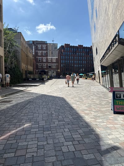 This is a street-level, vertical, full shot image taken in Groningen, Netherlands, on a sunny day. The scene depicts a cobblestone plaza or street lined with modern and older buildings.
On the left side of the image, a modern building with vertical wooden elements is visible, adjacent to a building with scaffolding and greenery. Further down the street, a multi-story brick building with numerous windows is present.
In the center of the image, several people are walking. Two men in the foreground are walking away from the camera, one in a pink t-shirt and the other in a light green t-shirt. A woman in a white top and jeans is walking in the same direction. Further back, a woman in a white top and a man in a light brown top are walking away from the camera.
On the right side of the image, a large, modern building with a textured concrete facade and a prominent sign that reads "FORUM GRONINGEN" is partially visible. A directional sign with the text "GRONINGEN" and "THIS WAY" in pink and green is positioned at the lower right corner.
The ground is paved with grey and pinkish cobblestones, with shadows cast by the buildings. The sky is blue with scattered white clouds. The overall impression is of a bustling urban environment on a pleasant day.