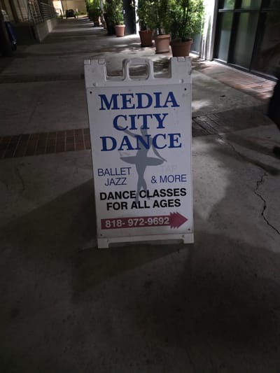 A white A-frame sign for "MEDIA CITY DANCE" stands on a cracked concrete sidewalk in front of a building. The sign features blue text and a silhouette of a dancer. Below the dancer, the sign lists "BALLET JAZZ TAP & MORE" and "DANCE CLASSES FOR ALL AGES". A phone number, "818-972-9692", is displayed in red at the bottom of the sign with a pink arrow pointing to the right. The building behind the sign has large windows and potted plants line the walkway. A dark car is visible in the distance. The lighting suggests it is dusk or nighttime.