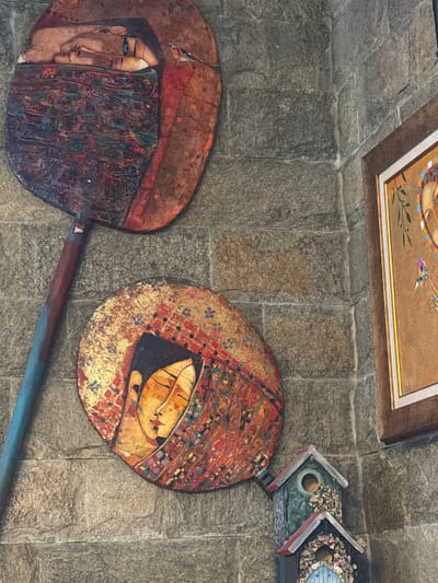 The image shows a section of a stone wall in what is possibly a public space, potentially in the city of Кирково, Bulgaria. Mounted on the wall are two decorative objects that appear to be artistic representations of baking peels, or pizza shovels. Each has a large, round, disc-shaped head and a long handle.
The left peel features a partial image of a woman's face, with visible eye and facial features, partially obscured by dark, patterned fabric. The right peel depicts a bowl-shaped object. Both peels have a textured, colorful finish.
Above the peels, there is a framed artwork and what appears to be a birdhouse or similar decorative structure, possibly made of wood. The wall is constructed of irregular, grey stones.