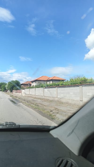 The image depicts a street in Boliartsi, Bulgaria, as seen from inside a vehicle. The road is paved with cracks and patches, and the view is obstructed by the car's dashboard and interior. On the right side of the road, a long, gray concrete block wall extends along the property line of a house with an orange tile roof. Vegetation is growing over the wall. In the distance, trees line the street. The sky above is partly cloudy, with blue visible.