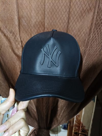 A black, faux leather baseball cap featuring a raised, monochromatic "NY" (New York Yankees) logo is centrally positioned in the frame, set against a draped, light brown fabric background. The cap has a structured crown and a flat brim with visible horizontal stitching lines. In the lower-left foreground, a hand, showing light to medium skin tone and natural nails, holds and partially fans out several banknotes, which display blue, pink, and cream colorations, consistent with Indonesian Rupiah. The background fabric exhibits a repeating diamond or lattice pattern. The scene is situated indoors, in Cileungsi, Indonesia.