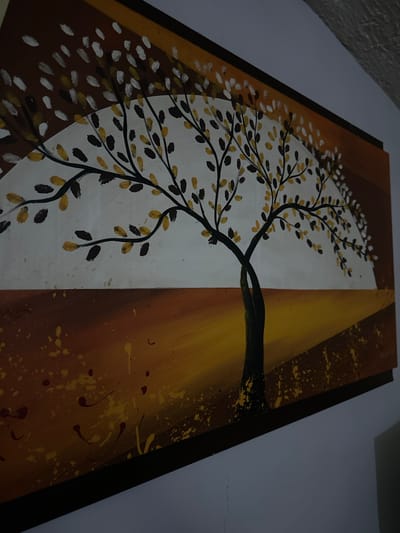 The image shows a rectangular painting with a dark brown frame, hanging on a light-colored wall. The painting is divided vertically into two distinct sections. The left section features a white background with a stylized tree. The tree has black branches adorned with leaves, rendered in a mix of gold and black. The leaves appear to be partially transparent. The right section has a brown background with yellow and darker brown splatters. A gradient of yellow is also visible. The tree trunk, depicted in dark brown and black, extends from the right section into the left. The photograph appears to be taken indoors under dim lighting conditions. There is no evidence to suggest the location is in Parroquia Ocumare, Venezuela.