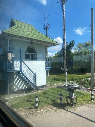 The image shows a two-story building with a green tiled roof, a light-colored facade, and blue metal railings. A tall metal tower with antenna-like structures rises from behind the building. A utility pole with horizontal supports and wires is visible in the upper left of the frame. A railroad crossing gate is partially visible in the upper right of the image, with black and white striped posts alongside a road. The sky is blue with some clouds. The location is Pidluky, Ukraine. The view is through a window, as evidenced by reflections in the foreground.