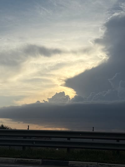The image shows a sky with dramatic cloud formations. On the left side, the clouds are light gray and white, with a texture resembling wisps or streaks. A golden light seems to be shining through parts of these clouds. On the right side, the clouds are dark gray, suggesting a storm or rain cloud. A road barrier appears in the right side of the image. The scene likely is in Чистенское сельское поселение, Ukraine, judging by the information provided.