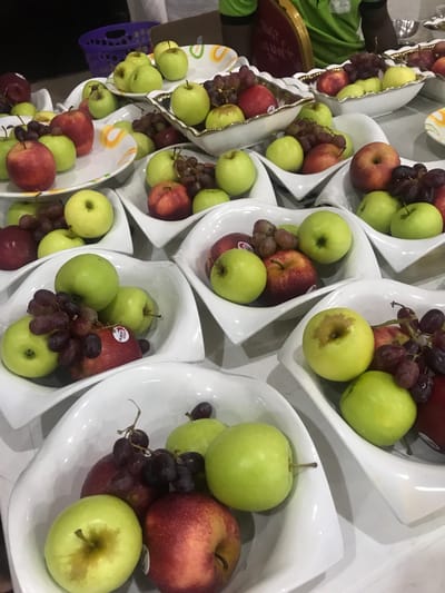 The image shows multiple white, modern-style bowls filled with green and red apples, and bunches of red grapes. The bowls are arranged in rows, with some placed on decorated tiered stands. A person wearing a green shirt can be seen in the background. The context indicates the location is likely in Ife, Nigeria.