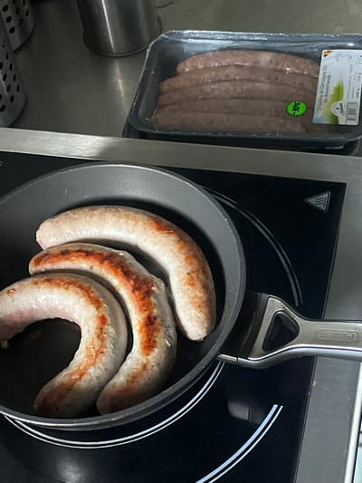 The image depicts a kitchen scene in Anderlecht, Belgium, featuring food preparation. In the foreground, a dark gray frying pan containing three partially cooked, light brown sausages with visible browning and char marks rests on a black induction cooktop. The cooktop displays white circular outlines indicating heating elements and a triangular 'hot surface' warning symbol. A metallic, silver-colored handle, branded "PYREX," extends from the pan to the right. In the midground, directly behind the pan, a black plastic tray sealed with clear film contains several uncooked, reddish-brown sausages. A green sticker with "$5" is affixed to this package, which also bears a label in French identifying the product as "Chipolata de Campagne Bio" (Organic Country Chipolata). The background consists of a reflective metallic surface, likely a kitchen counter or splashback, with indistinct metallic cylindrical objects visible to the upper left. No individuals are present in the image.