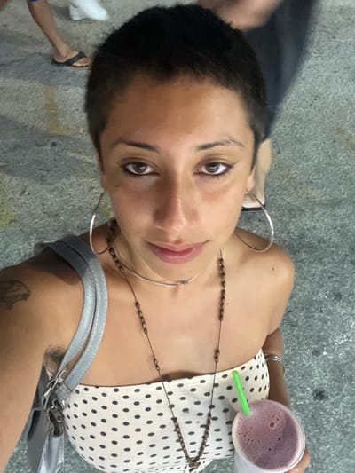 A woman poses in a close-up selfie outdoors. She has short dark hair, brown eyes, and wears large hoop earrings. A brown beaded necklace adorns her neck. She’s wearing a white polka dot top. A green straw is visible in a cup filled with a purple liquid that she holds near her shoulder. A silver purse strap is slung over her shoulder. Someone's feet are visible in the background. The background is pavement.