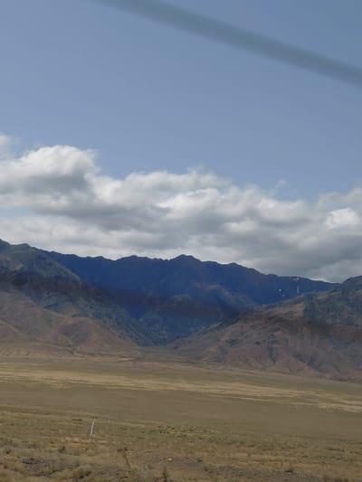 This image depicts a wide, open landscape dominated by mountains and a flat, grassy plain. In the foreground, the plain is covered in dry, yellowish-brown grass with sparse, darker vegetation. A tall, thin white post with a yellow band near the top stands in the middle ground, planted in the ground. Behind the plain, a range of mountains rises, with their lower slopes covered in brown and green vegetation. The upper peaks of the mountains are a deep blue, and some patches of snow or light-colored rock are visible on the higher elevations. The sky above is a pale blue, with scattered white and gray clouds. A dark, elongated shadow runs diagonally across the top of the image. The scene is characteristic of a rural or wilderness area. No people are visible in the image. The provided context suggests this location is in Ынталы, Kazakhstan, implying a steppe or mountainous terrain common to that region.