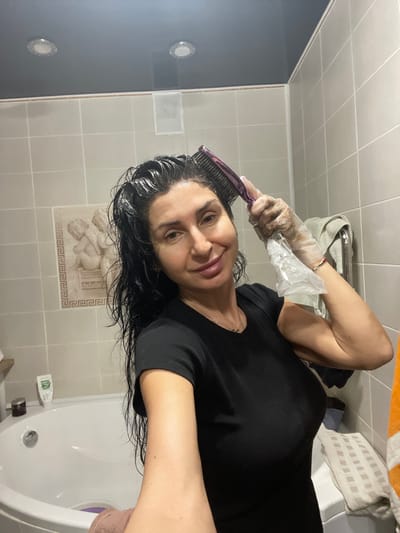 A woman in a black top applies hair dye with a purple comb while wearing a plastic glove. She is in a tiled bathroom with a white tub, and her wet hair is slicked back. Bathroom fixtures include a white vent and recessed lighting on the left side of the image. A towel and bag rest above the woman, near the upper edge. The woman is smiling and facing the camera.